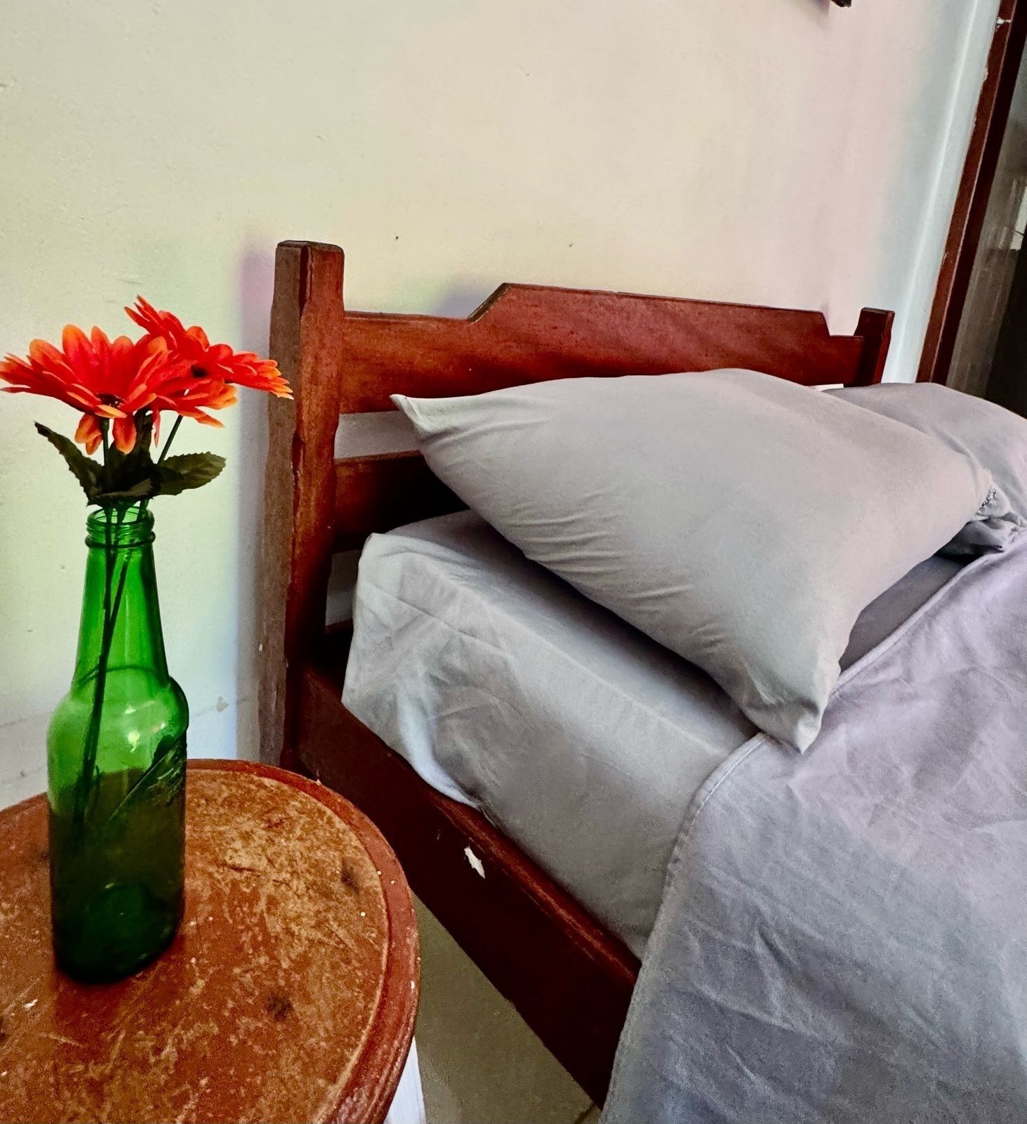 Een bed met kussens, een houten hoofdbord en een groene fles met bloemen op een ronde tafel.