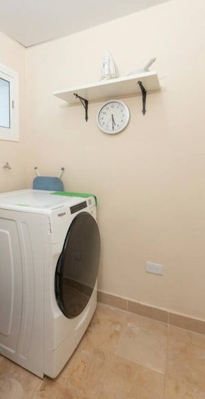 A laundry room with a front-loading washing machine, a clock, and a shelf with a small item on it. - Beachfront Condo | FREE Electricity | Sleeps 8 (Punta Cana)