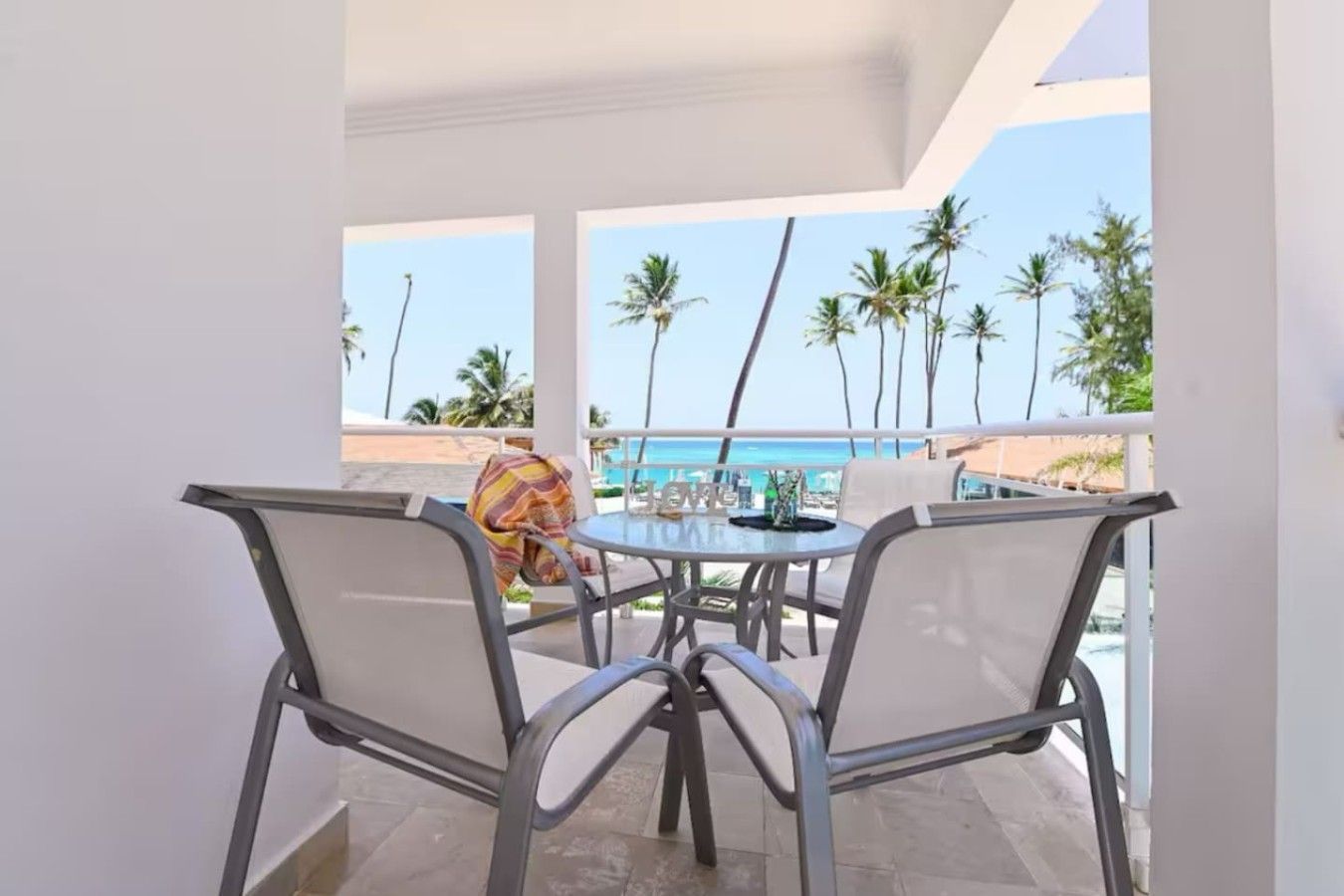 A balcony with a table and chairs, overlooking a beach with palm trees and the ocean.