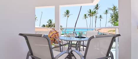 A balcony with a table and chairs, overlooking a beach with palm trees and the ocean.
