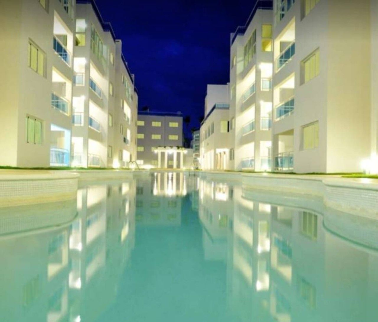 Une piscine dont l’eau reflète les bâtiments environnants, le tout étant situé dans un espace bien éclairé.