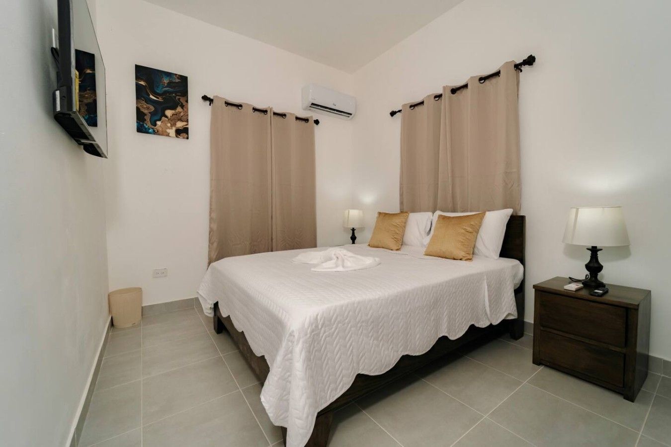 A bedroom with a bed, curtains, a bedside table with a lamp, a wall-mounted air conditioner, and a painting.