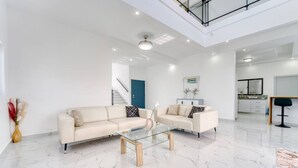 Een ruime woonkamer met een glazen salontafel, twee banken, een blauwe deur en een trap naar een hoger gelegen verdieping.