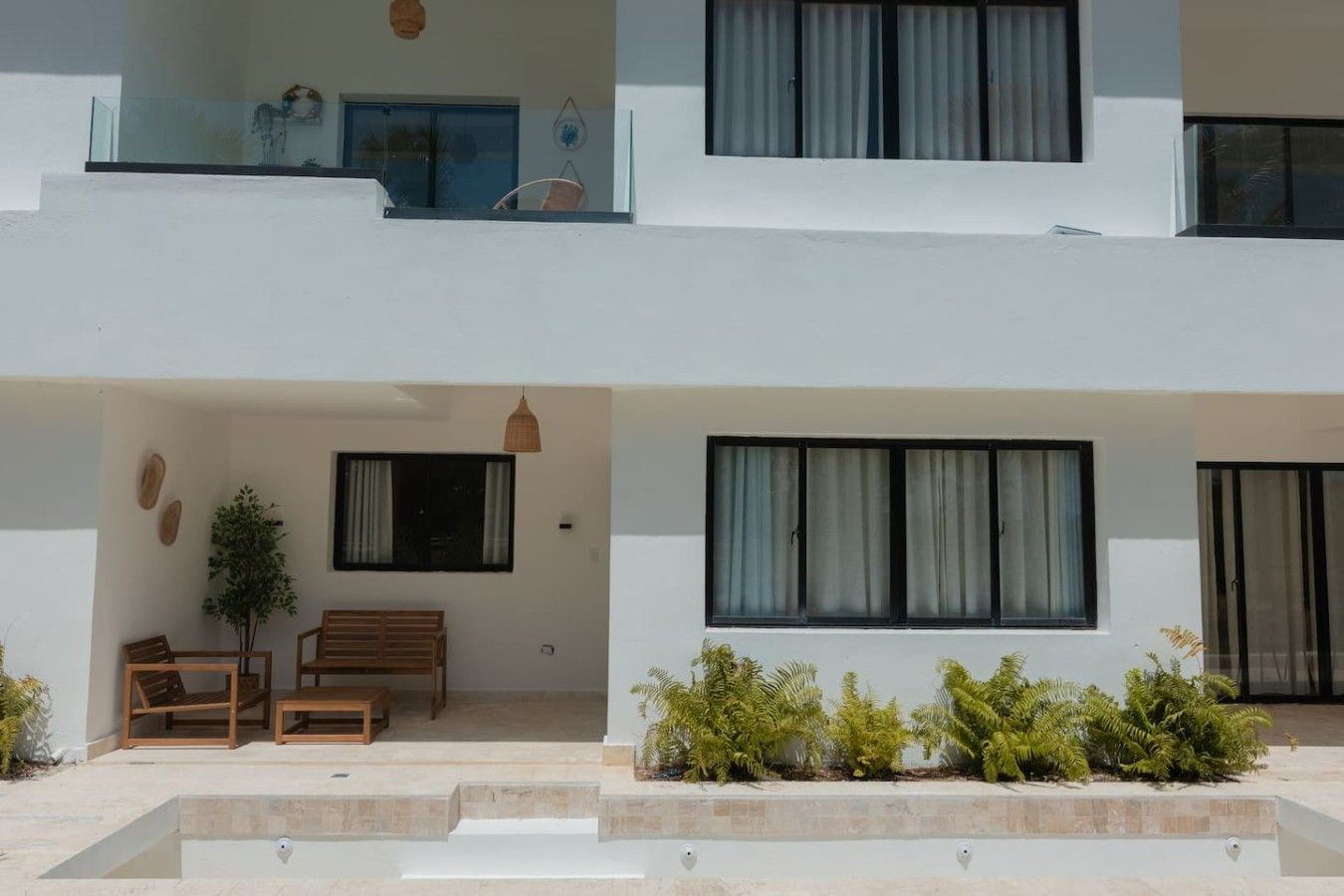 A modern two-story building with a balcony, a seating area with chairs and a table, and a small garden with plants.
