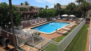 Ein Swimmingpool mit Liegestühlen und Sonnenschirmen, umgeben von einem eingezäunten Bereich und Palmen.