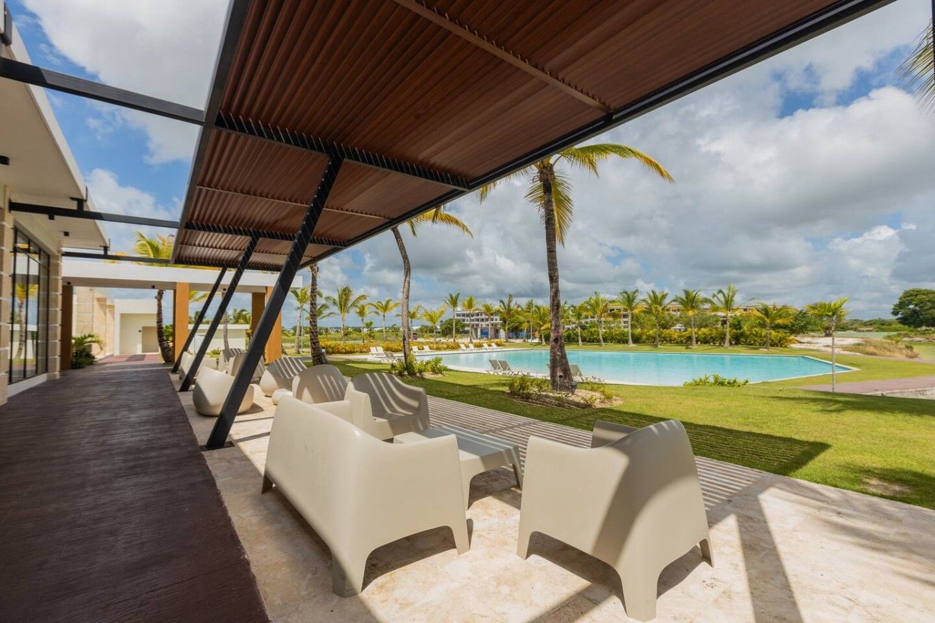 A poolside area with lounge chairs, a covered patio, and a view of a swimming pool and palm trees.