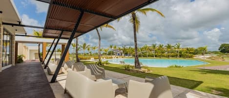 Zona junto a la piscina con sillas de descanso, un porche cubierto y vistas a una piscina y palmeras.