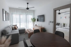 Ein modernes Wohnzimmer mit einer Couch, einem Couchtisch und einem Esstisch. Ein Fernseher ist an der Wand befestigt und ein Deckenventilator vorhanden.