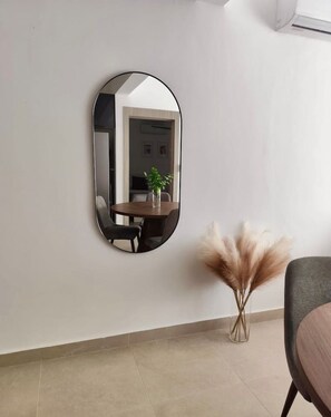 Ein Zimmer mit einem runden Spiegel an der Wand, einem Esstisch mit Stühlen, einer Vase mit Pampasgras und einem Sessel mit Kissen.
