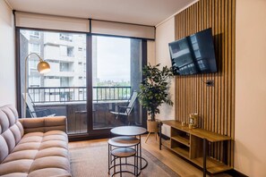 Ein modernes Wohnzimmer mit einer Couch, einem kleinen Tisch, einem an einer Holzwand montierten Fernseher und einem Balkon mit Blick auf Gebäude.