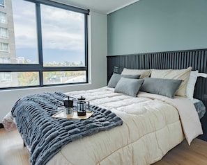 Ein modernes Schlafzimmer mit einem großen Fenster, einem Bett mit grauer Decke und einem Tablett mit Kaffeekanne und Tasse.