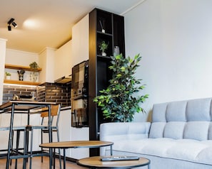 Ein modernes Wohnzimmer mit grauem Sofa, einem Esstisch mit Stühlen, einer Küchenzeile mit Backsteinwand und einer großen Grünpflanze.