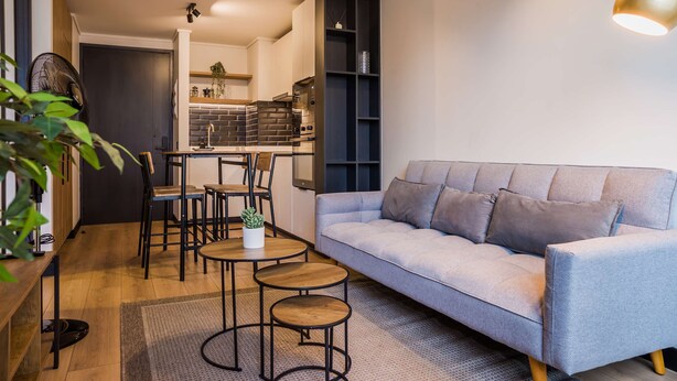 Un salon moderne avec un canapé gris, une petite table basse et un coin repas, la cuisine étant visible au fond.