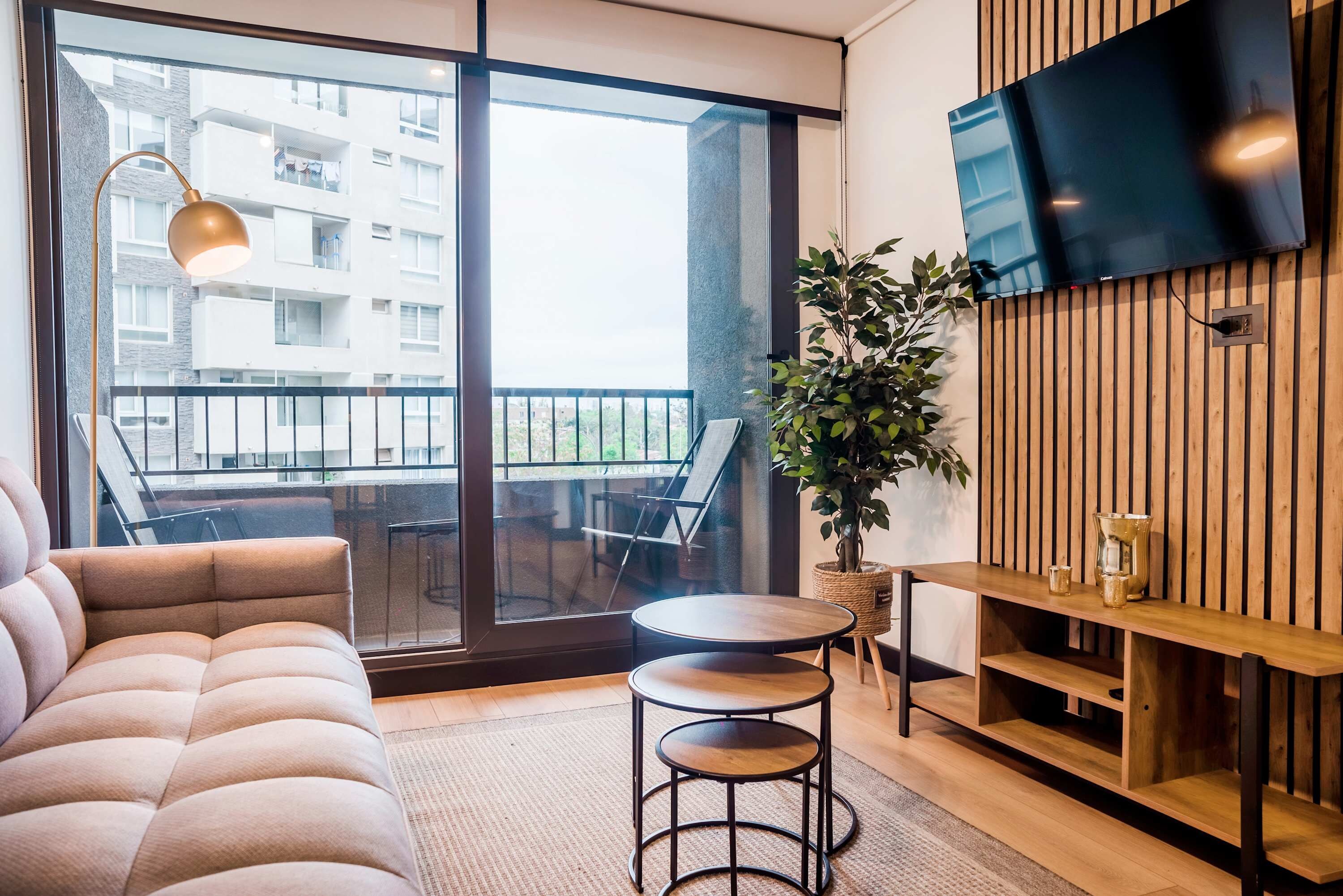 A modern living room with a sofa, a small round table, a potted plant, a wooden TV stand, and a balcony with a view of a building.