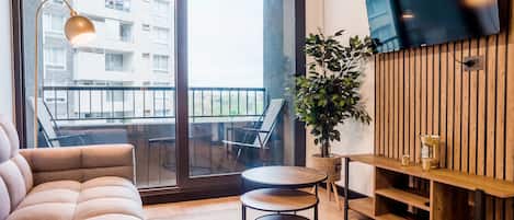 Ein modernes Wohnzimmer mit einer Couch, einem kleinen runden Tisch, einem Blumentopf, einer Holz-TV-Konsole und einem Balkon mit Blick auf ein Gebäude.