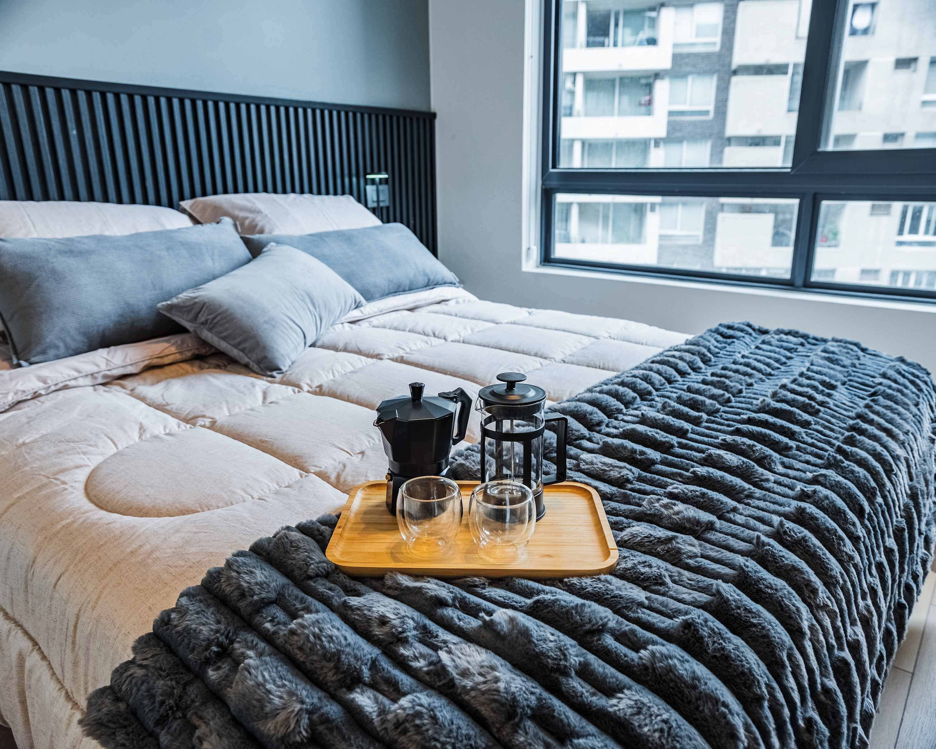 A neatly made bed with a grey blanket, a wooden tray with a coffee maker, two glasses, and a teapot.