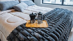 Ein ordentlich bezogenes Bett mit einer grauen Decke, ein Holztablett mit einer Kaffeemaschine, zwei Gläsern und einem Teekessel.