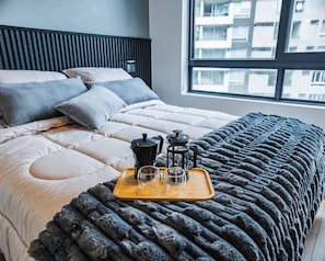 A neatly made bed with a grey blanket, a wooden tray with a coffee maker, two glasses, and a teapot.