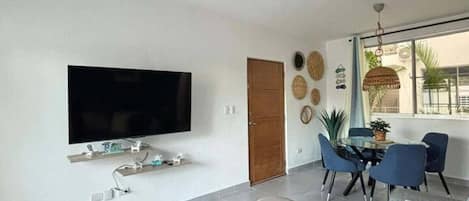 A modern living room with a ceiling fan, a flat-screen TV, a dining area with blue chairs, and a large window.