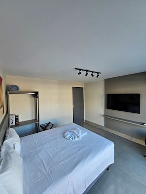 Et hotelværelse med en seng, et fjernsyn ophængt på væggen, en dør og en hylde med genstande.