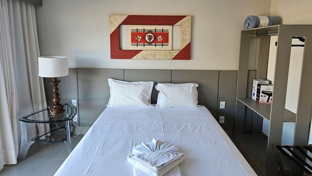 Quarto de hotel com cama bem arrumada, obra de arte na parede, luminária de cabeceira e pequena mesa com abajur.