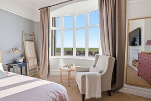 Ein Schlafzimmer mit großem Fenster, einem Bett, einem Sessel, einem Schminktisch und einer Lampe.