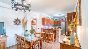 A dining area with a glass table, wooden chairs, a chandelier, and a kitchen in the background. - Cubo's Marala Rural House / Private pool / BBQ / Terrace / Free Wi-Fi (Alhaurín el Grande)