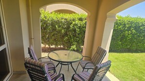 A patio with a round glass table and four striped chairs, surrounded by a low wall and a hedge.