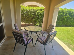 Eine Terrasse mit einem runden Glastisch und vier gestreiften Stühlen, umgeben von einer niedrigen Mauer und einem Hecken.