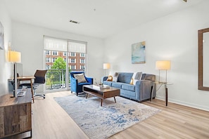 Ein modernes Wohnzimmer mit Sofa, Sessel, Couchtisch und Blick auf eine Stadtlandschaft durch große Fenster.