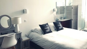 A hotel room with a bed, a desk with a lamp, a chair, a mirror, a bathroom with a shower, and a sink. - Hôtel Le Galet Bleu (Fouras)