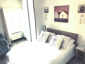 A bed with white bedding and two pillows, a bedside table with a lamp, a framed picture of a house, and a window with curtains. - Hôtel Le Galet Bleu (Fouras)