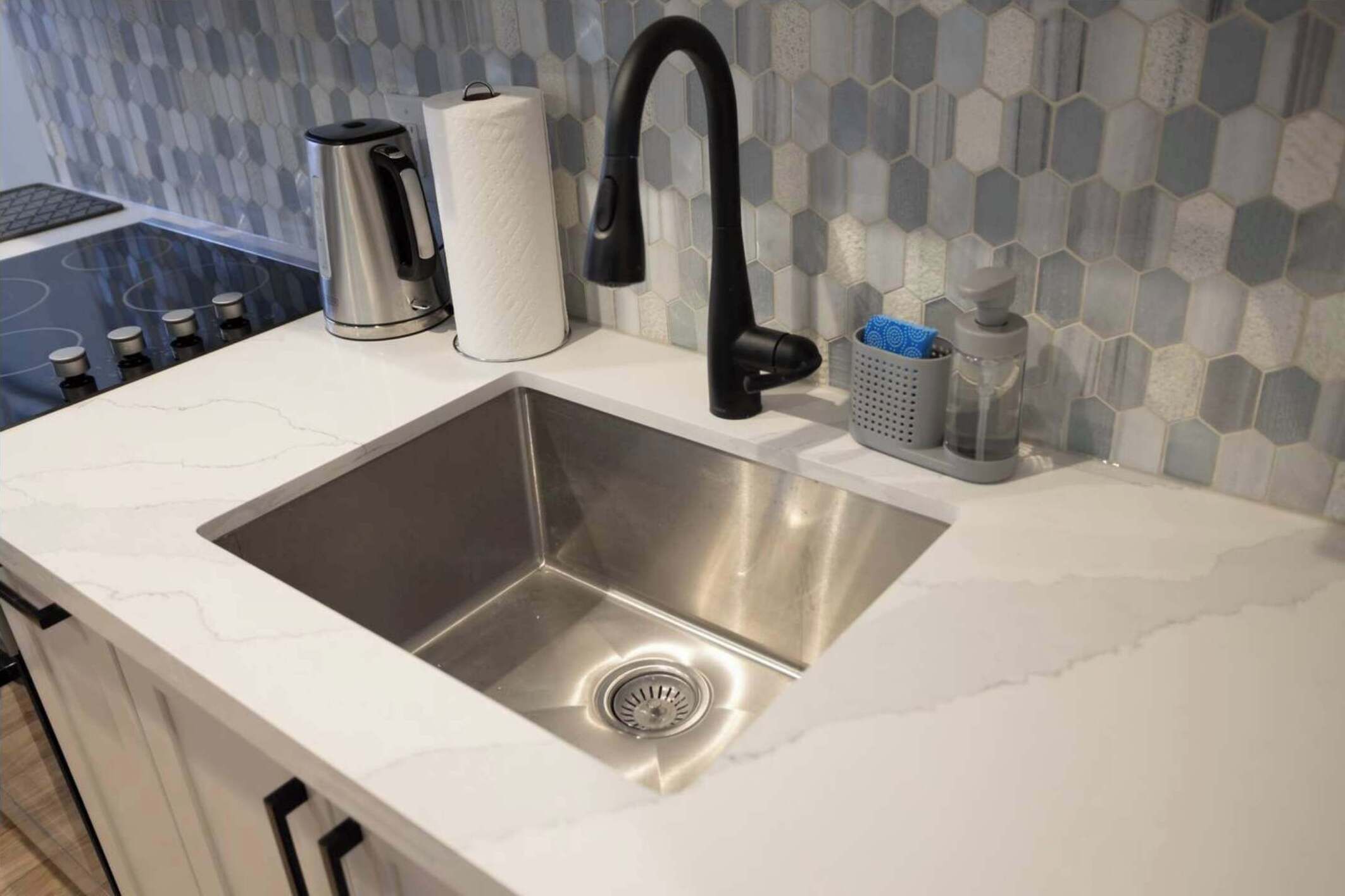 Ett kök med en diskho i rostfritt stål, en svart blandare, en vattenkokare och en hållare för hushållspapper.