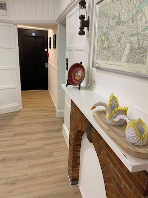 Un couloir doté d’un plancher en bois, d’un mur blanc orné d’une carte encadrée, d’une assiette décorative rouge et de cygnes en papier posés sur un manteau de cheminée blanc.