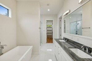 Ein modernes Badezimmer mit einer Badewanne, zwei Waschbecken, einem großen Spiegel und Blick auf eine grüne Außenanlage.