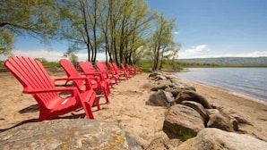 A row of red chairs lined up on a sandy beach with rocks, trees, and a body of water in the background.