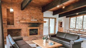 A living room with a brick fireplace, leather sofas, a wooden coffee table, and a view of trees through large windows.