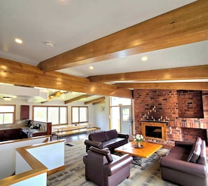 A spacious living area with a fireplace, wooden beams, and a kitchen in the background. - 6 bdrm Cottage, Private Beach Access, HotTub/Sauna (The Blue Mountains)