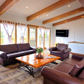 A living room with a wooden table, leather sofas, a flat-screen TV, and large windows with curtains. - 6 bdrm Cottage, Private Beach Access, HotTub/Sauna (The Blue Mountains)