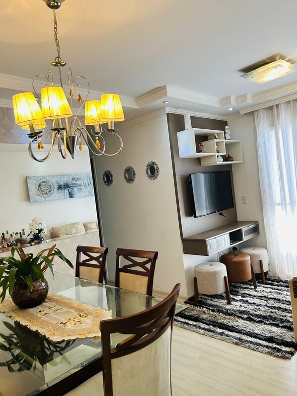 Un salon moderne avec une table à manger en verre, des chaises en bois, un lustre, un téléviseur à écran plat et une grande fenêtre ornée de rideaux légers.