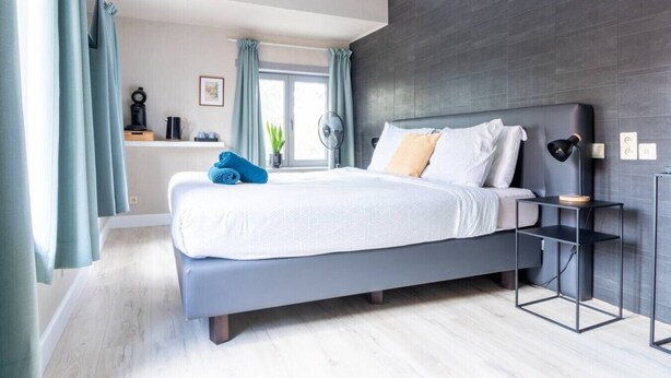 Une chambre moderne avec un lit gris, une table de chevet avec une lampe, une fenêtre avec des rideaux et un mur foncé.