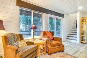 A cozy living room with a sofa, armchair, and a lamp. There are large windows with a view of trees.