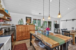 A kitchen with wooden cabinets, a dining table, hanging pendant lights, and a view of the outdoors.