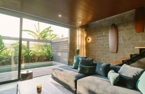 A modern living room with a large glass door leading to a pool, a grey sofa, and a wooden staircase. - Luxury 3BR Townhouse (Badung)