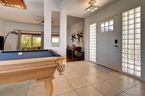 Ein Billardtisch in einem Raum mit Deckenventilator, einer Lampe und Blick auf einen Garten durch ein Fenster.