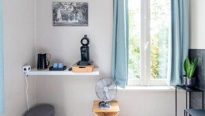Una stanza con una finestra, una foto in bianco e nero appesa al muro, una macchina da caffè, una ventola e una pianta.
