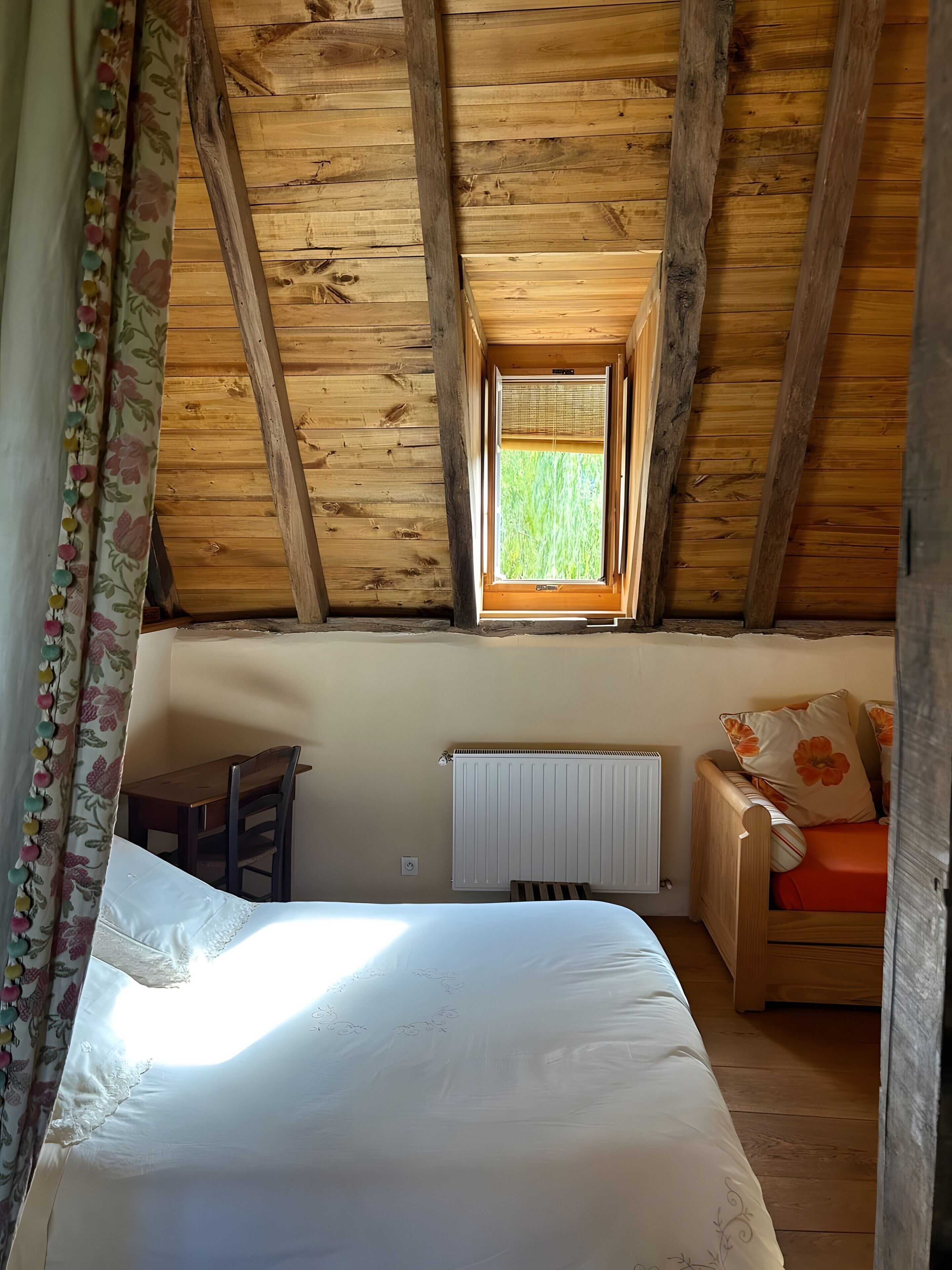 A room with a wooden ceiling, a bed, a small table, a radiator, and a window with a view of greenery.