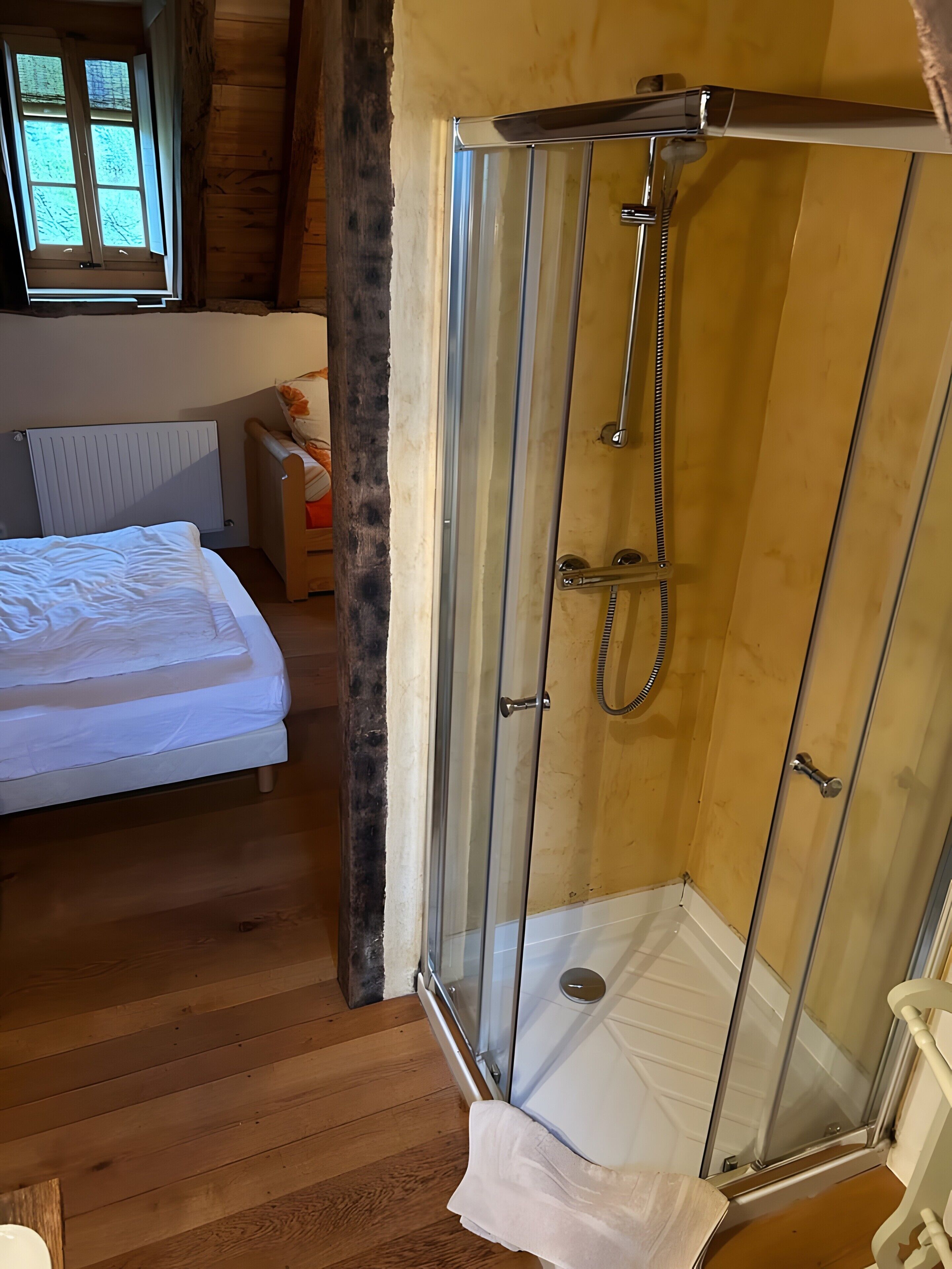 A room with a bed, a shower with glass doors, wooden flooring, and a window with curtains.