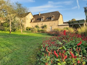 A stone building with a sloped roof, a grassy lawn, and red flowering plants in the foreground.