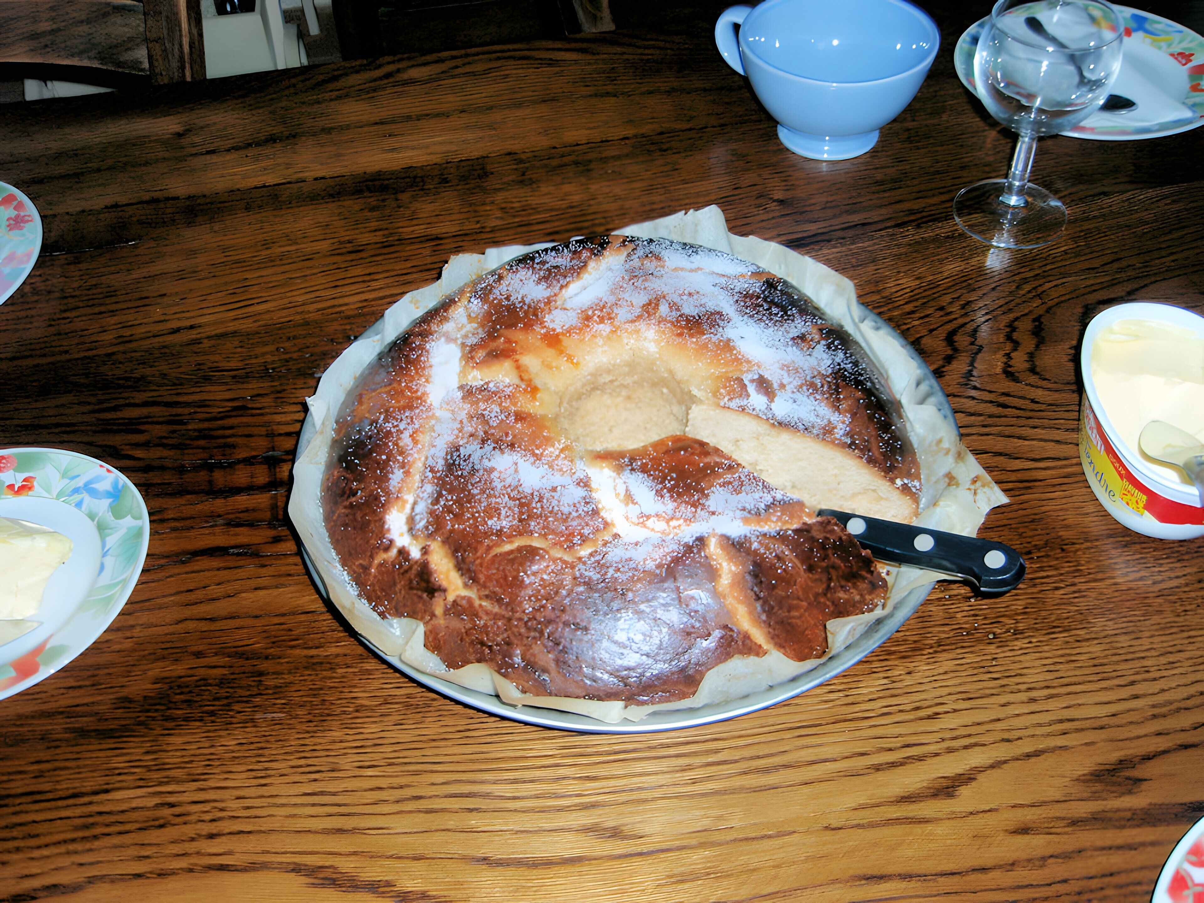 Ein runder Kuchen auf einem Teller mit einem Messer, einer blauen Tasse und einem Glas auf einem Holztisch.
